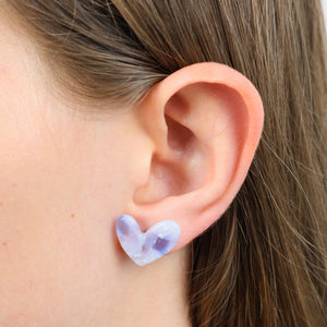 A close-up of a person's ear wearing a small heart-shaped stud earring in a pale lavender and white marbled stone or resin.