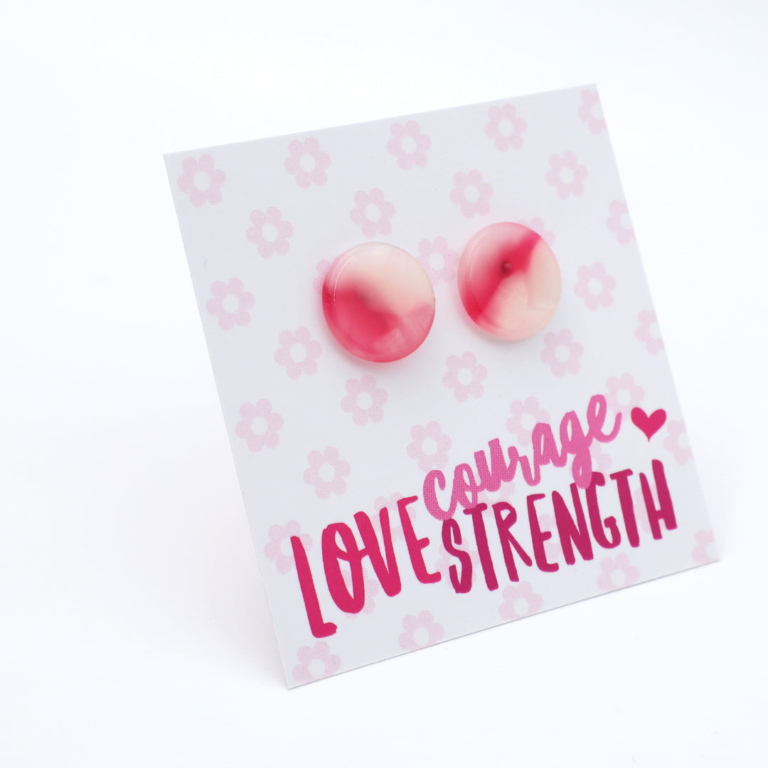 A pair of round pink and white marbled stud earrings displayed on a white backing card with a light pink floral pattern and the words 'courage LOVE STRENGTH' in pink lettering.
