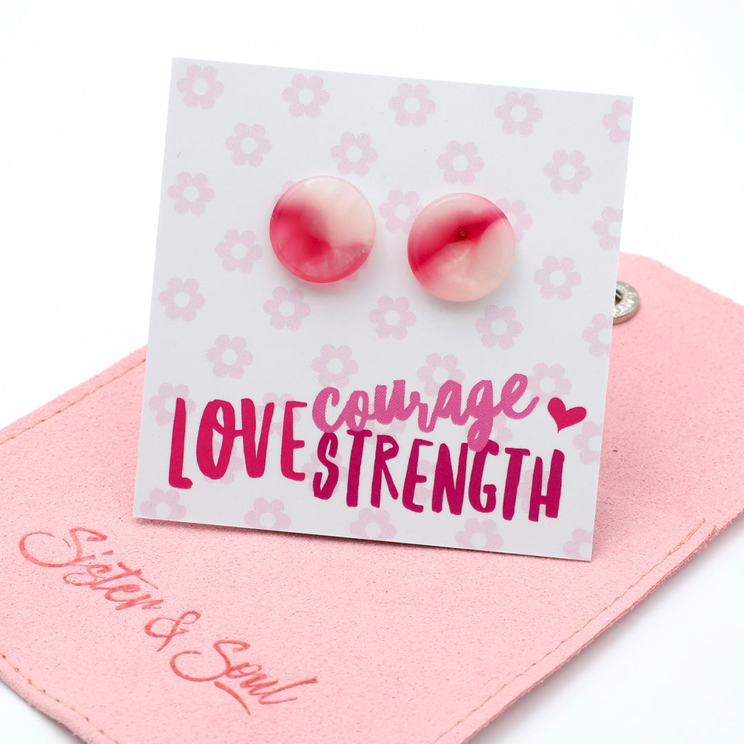 A pair of round stud earrings with a pink and white marbled pattern, displayed on a white backing card with the words 'LOVE STRENGTH' and 'courage' in script. The card is resting on a pink fabric pouch embroidered with 'Sister & Soul'.