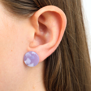 A close-up of a person's ear wearing a round lavender purple stud earring with a white marbled pattern.