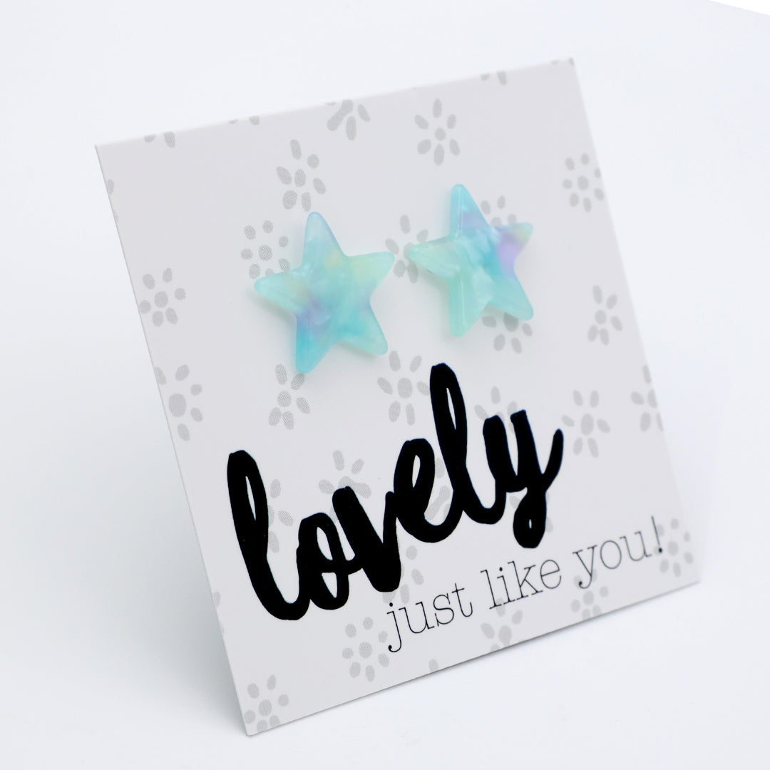A pair of star-shaped stud earrings in a light blue and lavender iridescent resin, displayed on a white printed card that reads 'lovely just like you!'.
