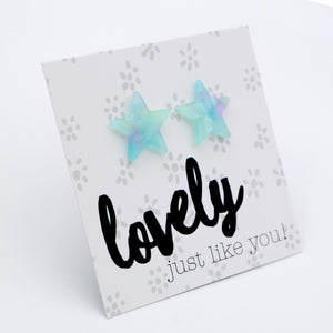 A pair of star-shaped stud earrings in a light blue and lavender iridescent resin, displayed on a white printed card that reads 'lovely just like you!'.