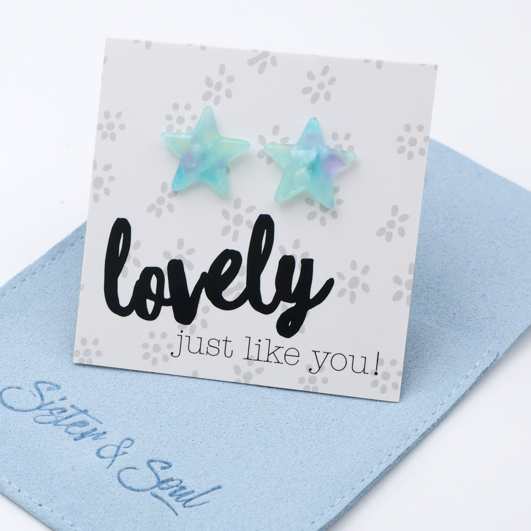 Two iridescent star-shaped stud earrings displayed on a white card that reads 'lovely just like you!' with a blue fabric pouch underneath. The earrings have a pastel blue and mint green color with subtle purple iridescence.
