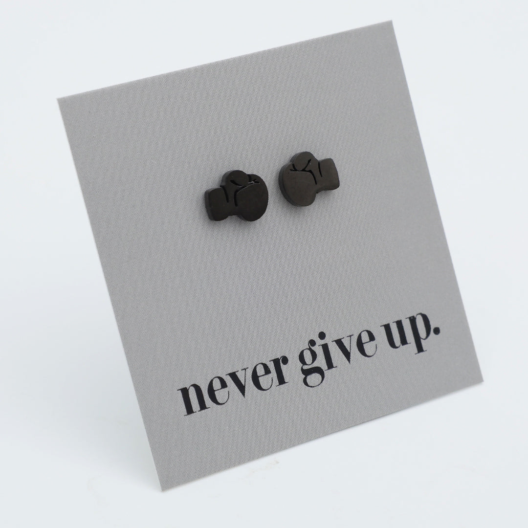 A pair of black stud earrings shaped like boxing gloves, mounted on a textured gray display card that reads 'never give up.' in black lowercase serif text, photographed on a clean white background.