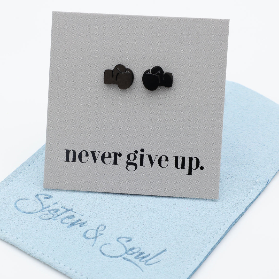 A pair of small black boxing gloves stud earrings displayed on a light gray card that reads 'never give up.' with a light blue fabric pouch underneath.