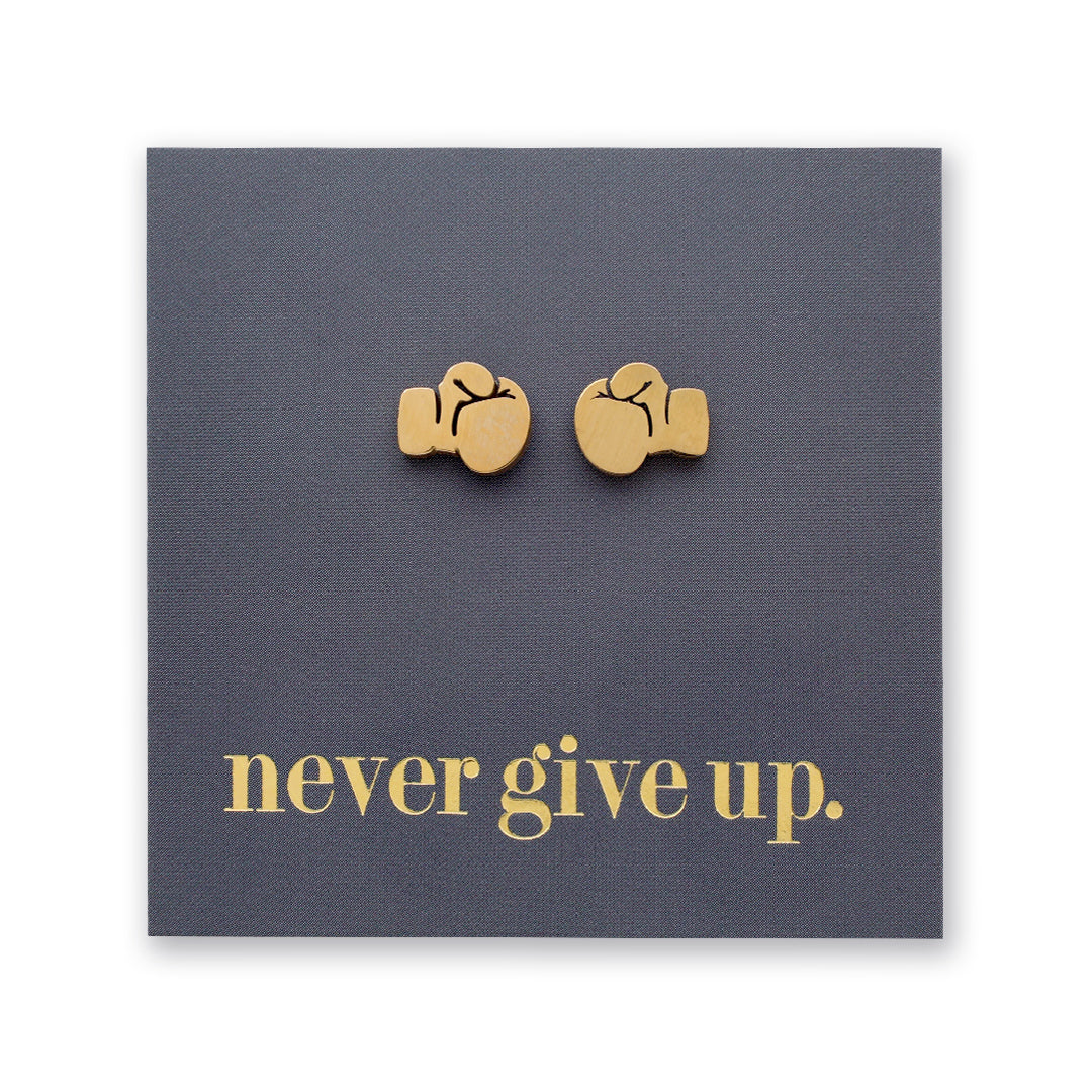 A pair of small gold stud earrings shaped like boxing gloves, displayed on a dark gray square backing card with the phrase "never give up." in gold lowercase text.