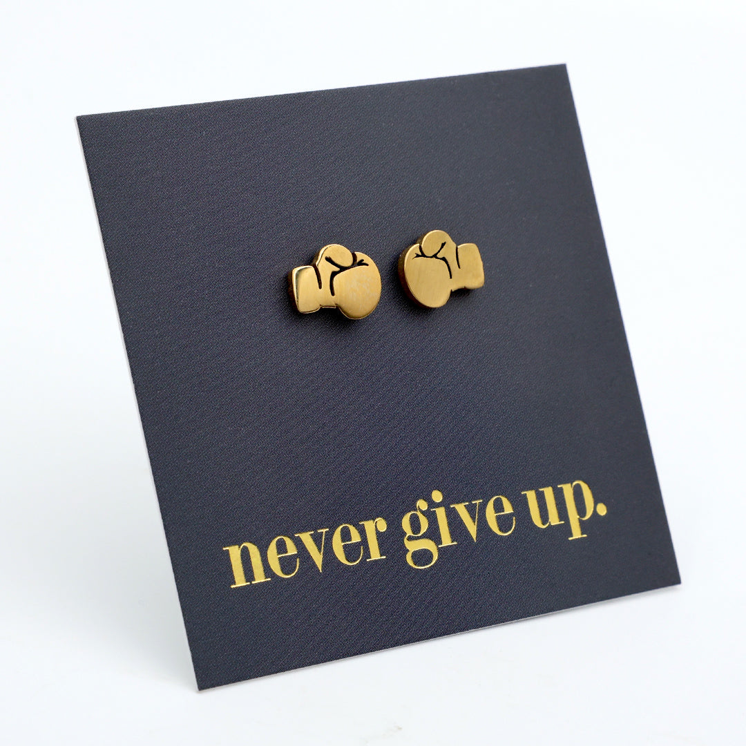 A pair of gold boxing glove stud earrings displayed on a black textured backing card with the phrase 'never give up.' in gold lettering, set against a clean white background.