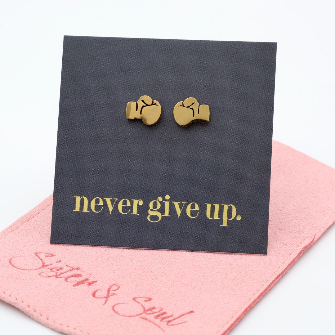A pair of small gold stud earrings shaped like b, displayed on a black backing card that reads 'never give up.' in gold lettering, with a pink fabric underneath pouch xing gloves