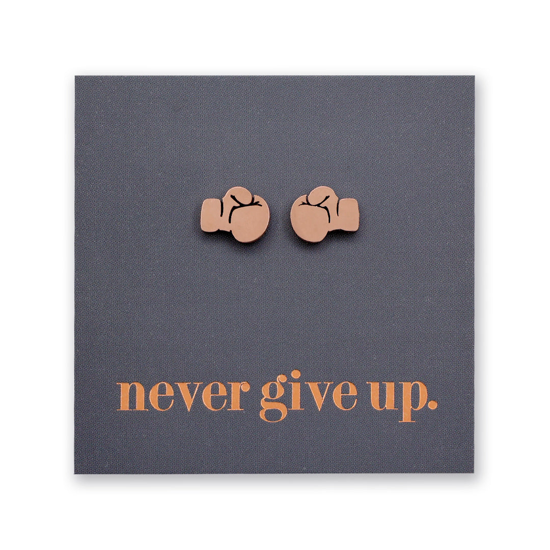 A pair of small rose gold stud earrings shaped like boxing gloves, mounted on a dark gray square backing card with the phrase 'never give up.' in rose gold  lettering.