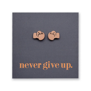 A pair of small rose gold stud earrings shaped like boxing gloves, mounted on a dark gray square backing card with the phrase 'never give up.' in rose gold  lettering.