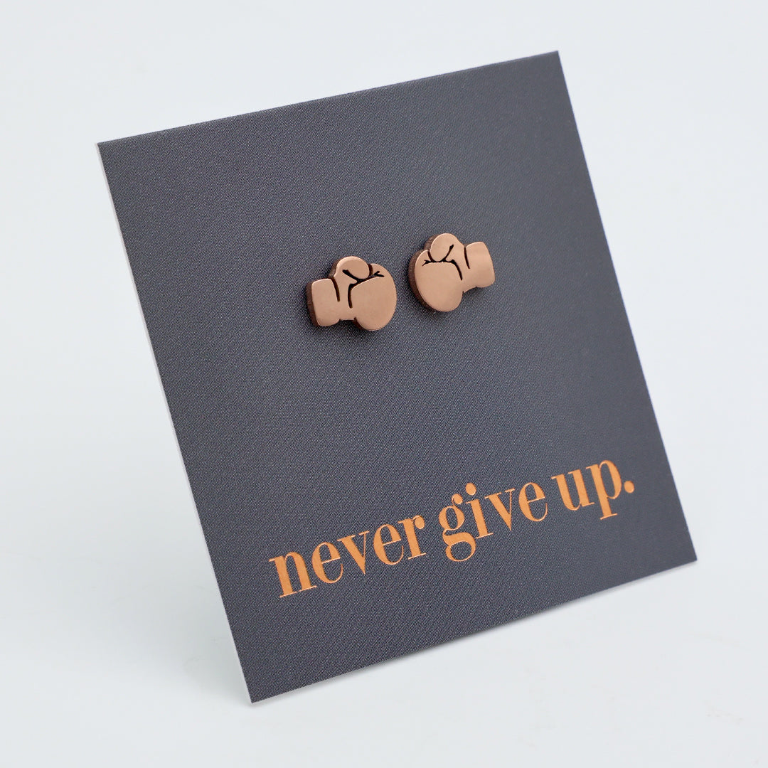 A pair of small rose gold stud earrings shaped like boxing gloves, mounted on a dark gray square display card that reads 'never give up.' in rose gold  text, photographed on a light gray background.