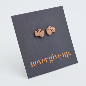 A pair of small rose gold stud earrings shaped like boxing gloves, mounted on a dark gray square display card that reads 'never give up.' in rose gold  text, photographed on a light gray background.