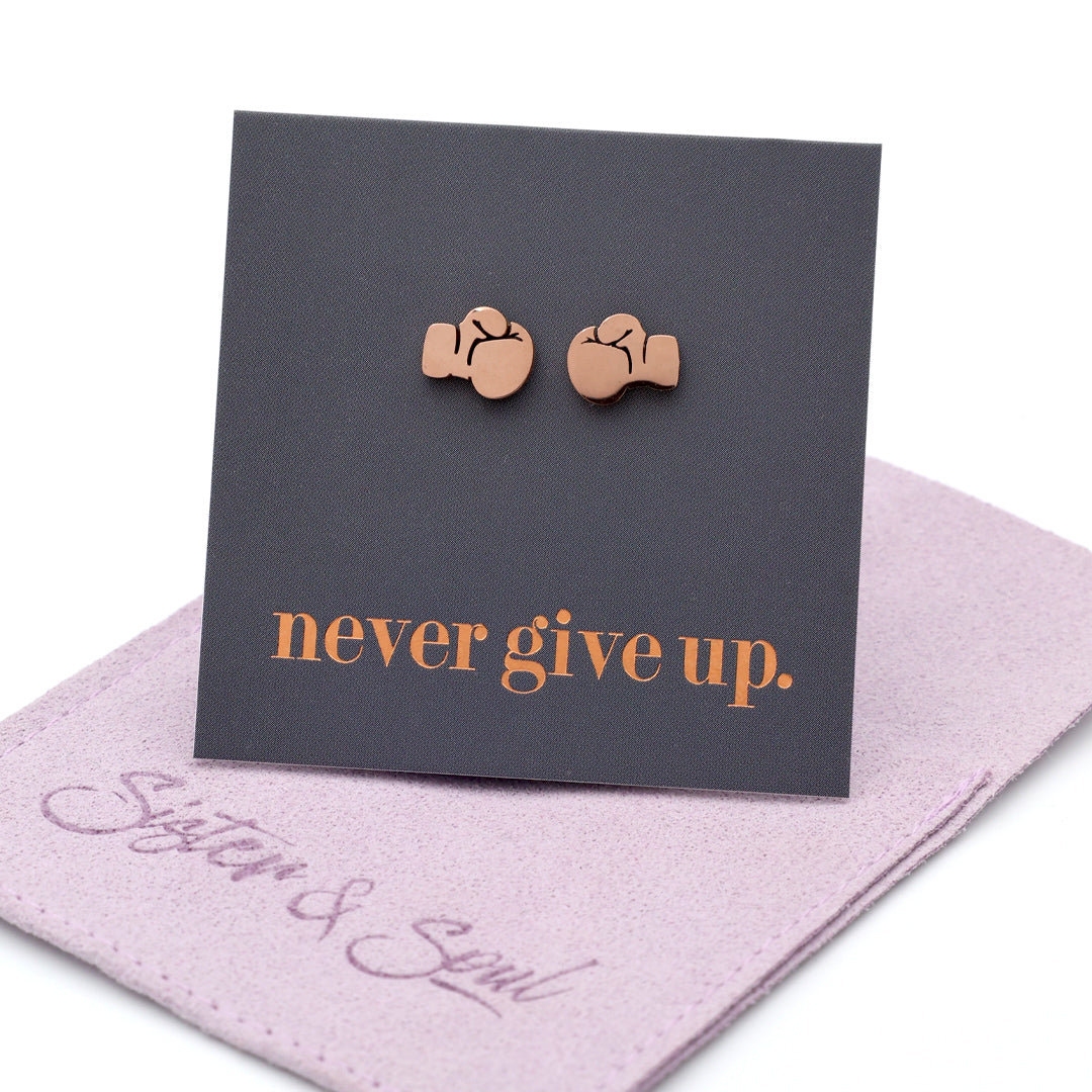 A pair of small rose gold stud earrings shaped like boxing gloves, displayed on a dark charcoal card that reads 'never give up.' in rose gold lettering, resting on a purple fabric pouch with the brand name 'Sister & Soul' visible.