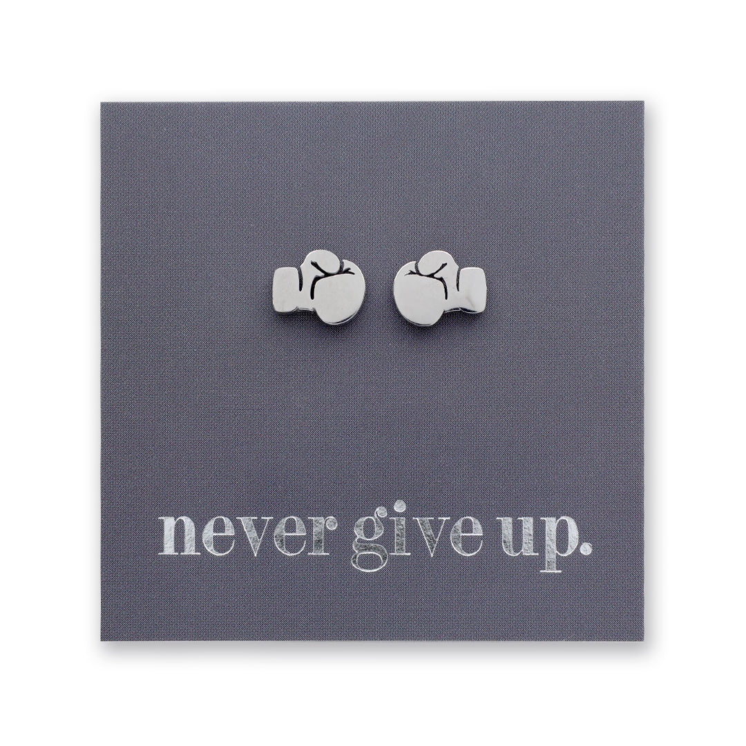 A pair of small silver stud earrings shaped like boxing gloves displayed on a square dark gray backing card that reads 'never give up.' in white lowercase text.