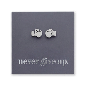 A pair of small silver stud earrings shaped like boxing gloves displayed on a square dark gray backing card that reads 'never give up.' in white lowercase text.