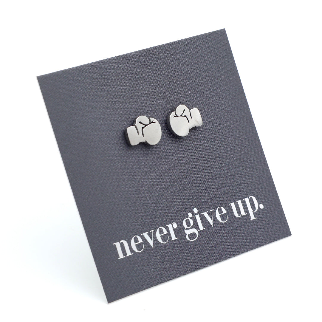 A pair of small silver stud earrings shaped like boxing gloves, displayed on a dark gray backing card that reads 'never give up.' on a white background.