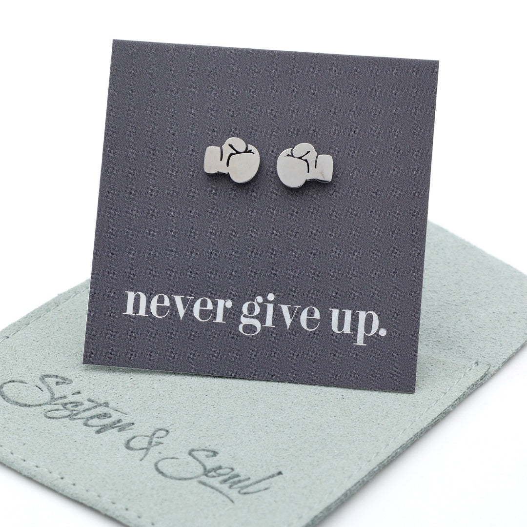 A pair of silver-toned stud earrings shaped like boxing gloves, displayed on a dark gray backing card that reads 'never give up.' with a light sage green jewelry pouch underneath on a white background.