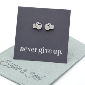 A pair of silver-toned stud earrings shaped like boxing gloves, displayed on a dark gray backing card that reads 'never give up.' with a light sage green jewelry pouch underneath on a white background.