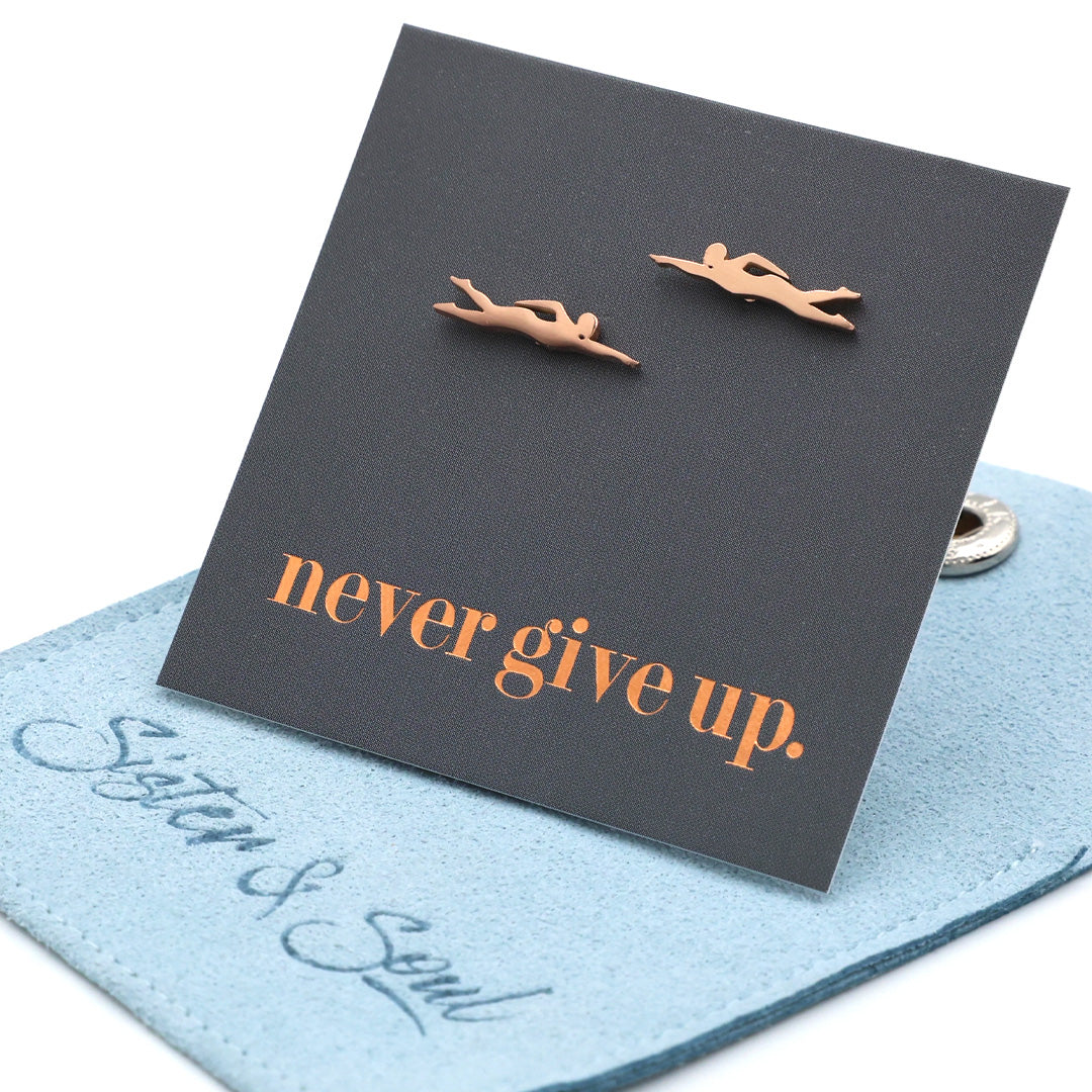 Rose gold swimming earrings on a black card with 'never give up.' text, placed on a blue cloth.