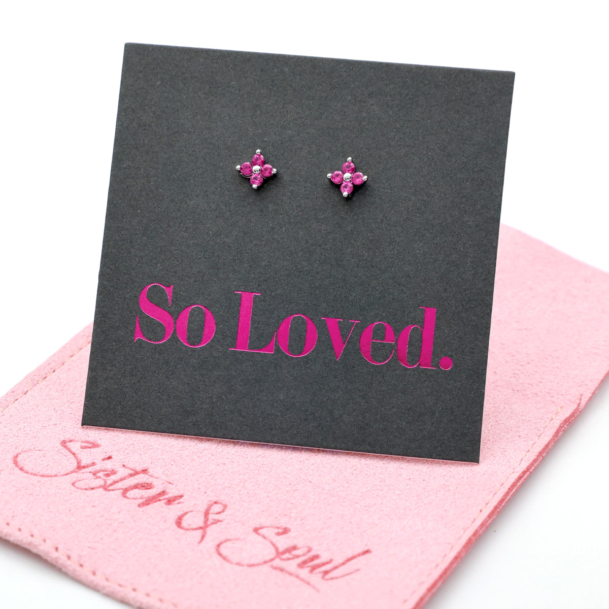 Pink tiny floral star shaped studs presented on a grey card with pink 'So Loved' writing sitting on a pink pouch with 'Sister & Soul' writing.