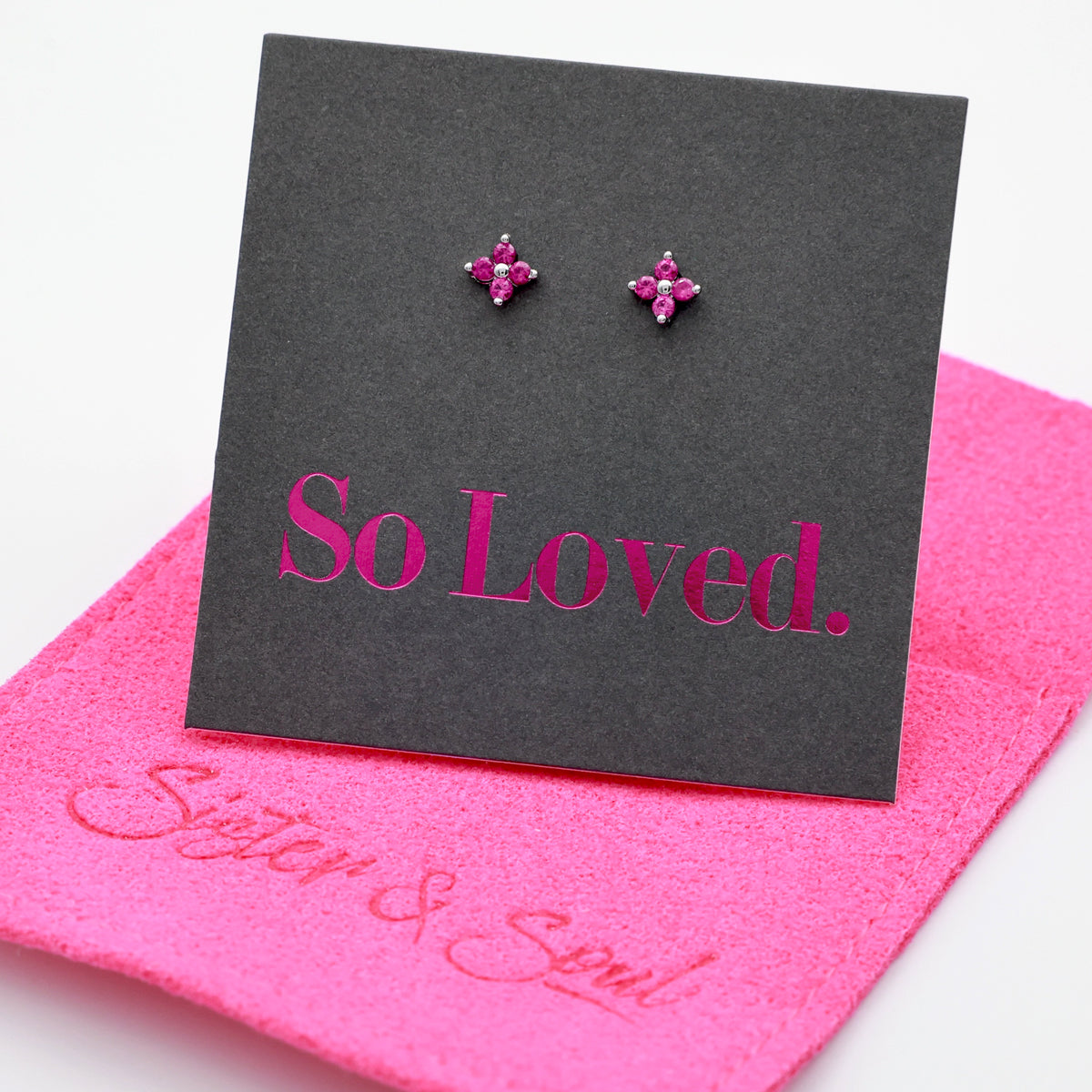 Pink tiny floral star shaped studs presented on a grey card with pink 'So Loved' writing sitting on a hot pink pouch with 'Sister & Soul' writing.