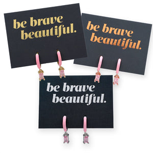 Black cards with 'be brave beautiful.' text and pink earrings on a white background