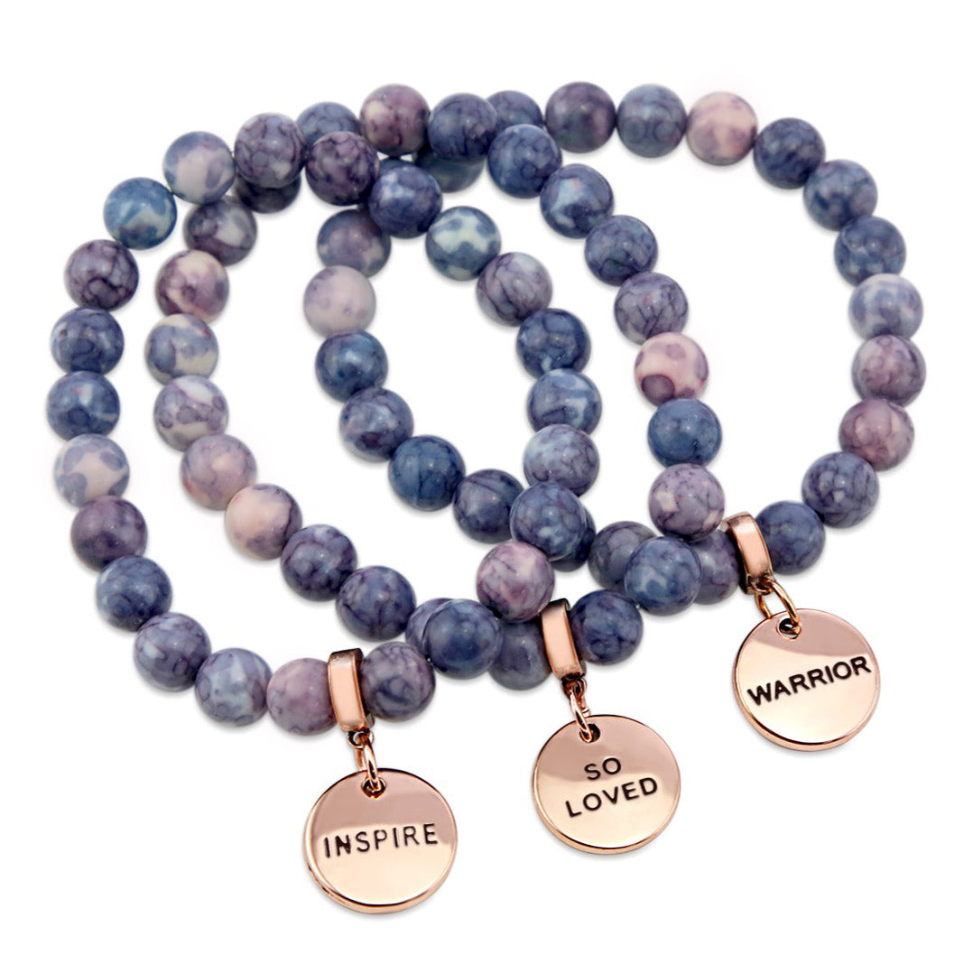 Two bracelets with purple and storm patch agate stone beads and rose gold word charms, one with 'INSPIRE' and the other with 'WARRIOR'.