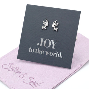 Silver reindeer earrings on a card with 'JOY to the world.' text, placed on a pink fabric surface with 'Sister & Soul' branding.