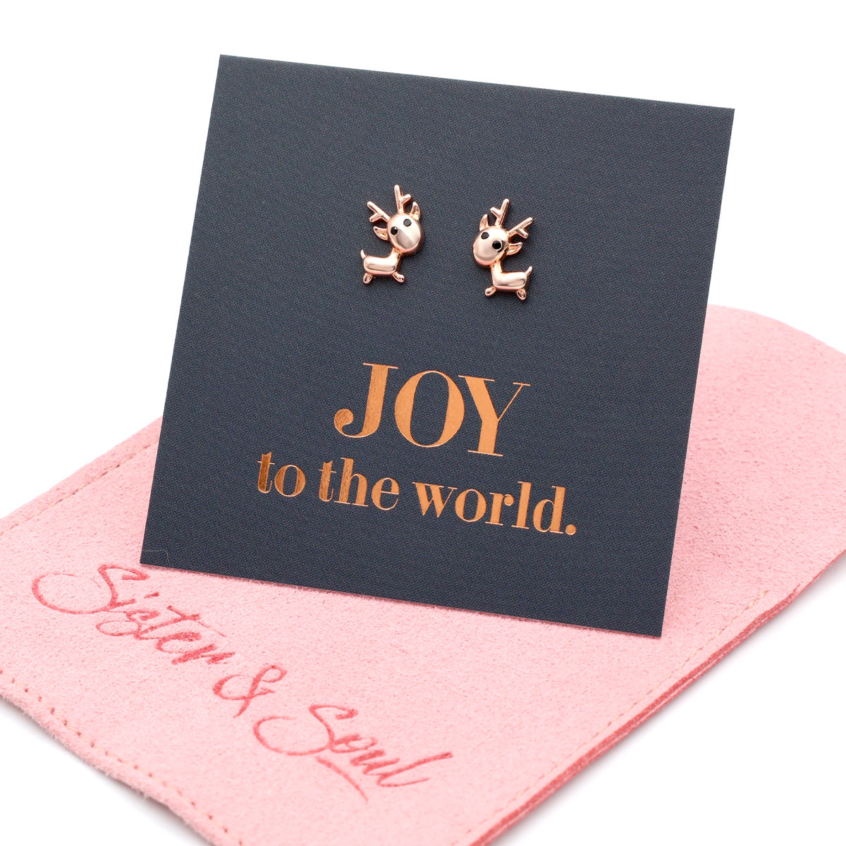 Rose gold reindeer earrings on a card with 'JOY to the world' text, placed on a pink cloth with 'Sister & Swirl' branding.