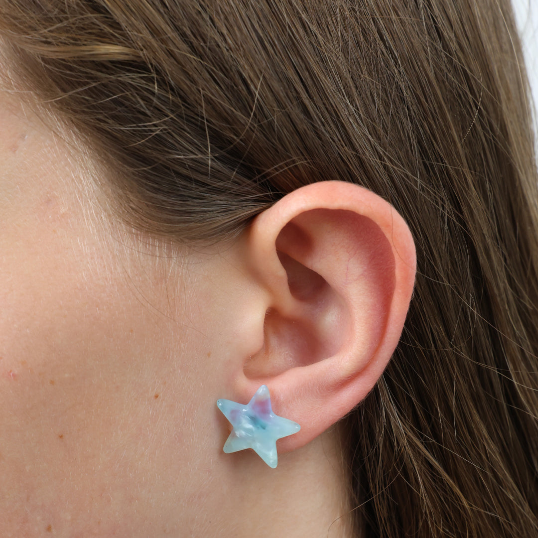 A pair of star-shaped stud earrings with a light blue and purple iridescent finish, displayed on a white square card that reads 'lovely just like you!' in black script.