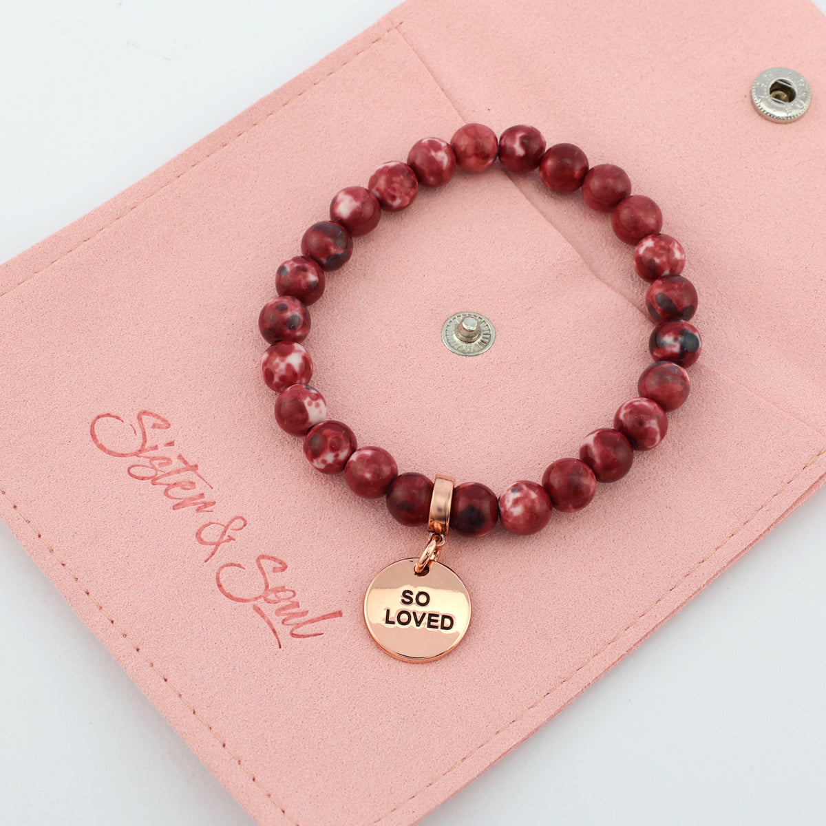Red beaded bracelet with a gold 'So Loved' charm on a pink card with 'Sister & Soul' branding.