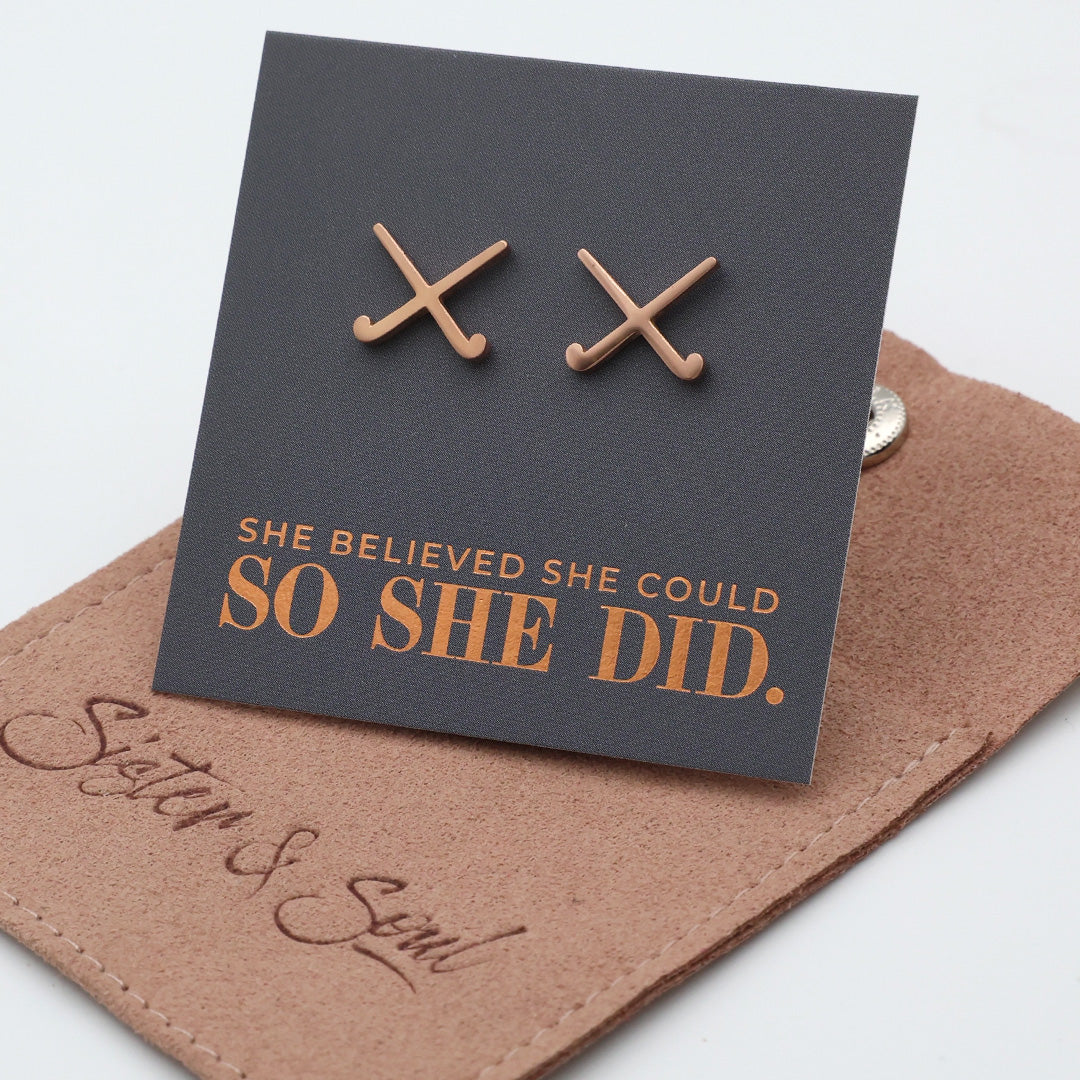 Rose gold hockey shaped earrings on a card with 'She believed she could so she did' text, on a brown surface with 'Sister & Soul' branding.