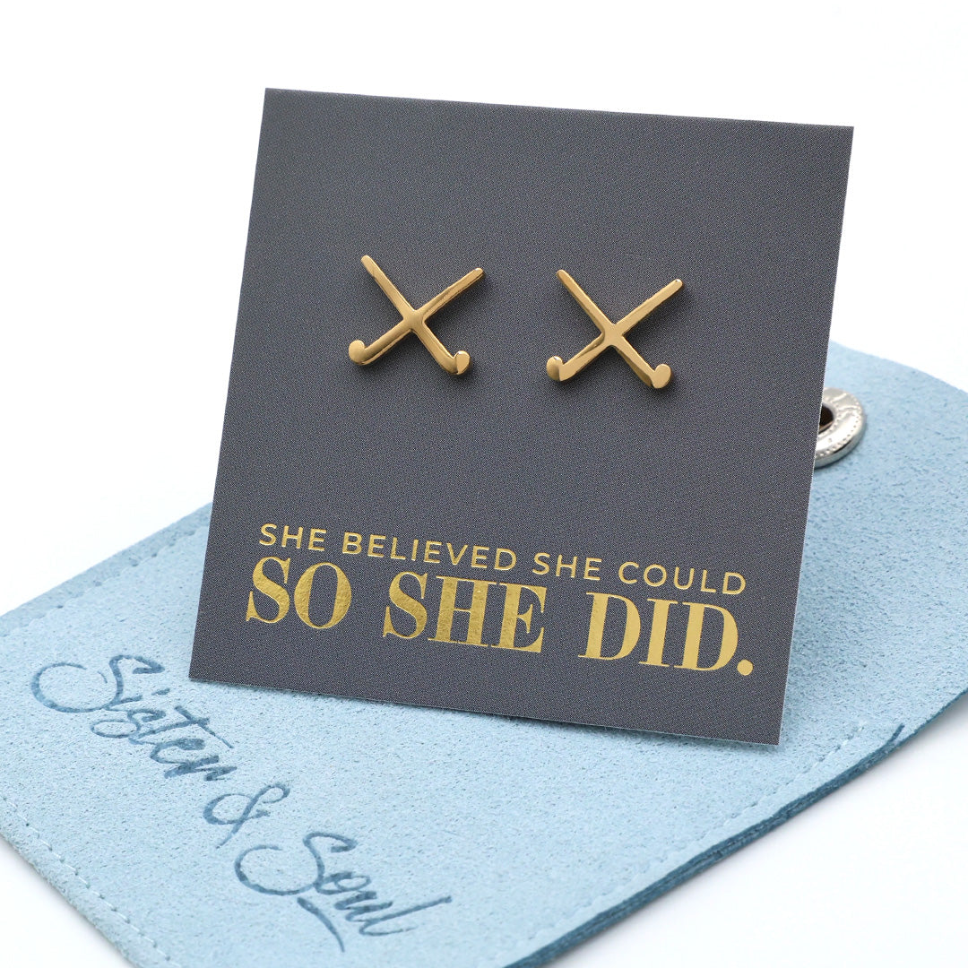 Gold stainless steel field hockey earrings on a card with 'She believed she could so she did' text, on a grey surface.