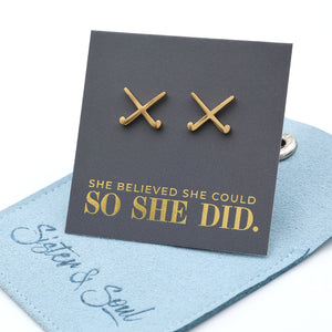 Gold stainless steel field hockey earrings on a card with 'She believed she could so she did' text, on a grey surface.