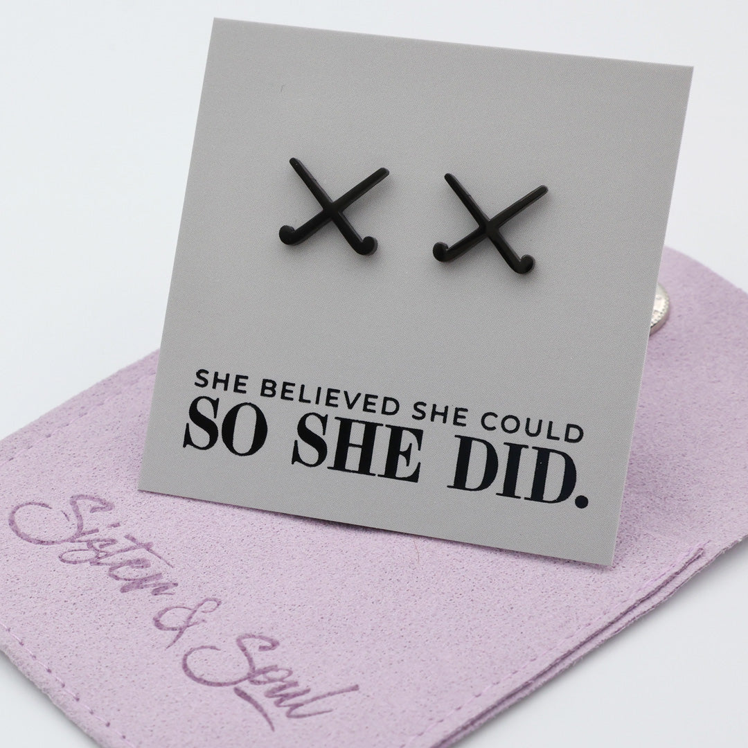Black stainless steel hockey-shaped earrings on a card with 'She believed she could so she did' text, displayed on a purple gift pouch.