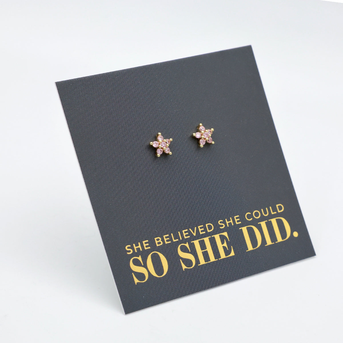 Pink cubic zirconia star shaped studs presented on a grey card with gold 'She Believed She Could So She Did' writing.
