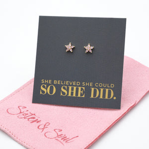 Pink cubic zirconia star shaped studs presented on a grey card with gold 'She Believed She Could So She Did' writing sitting on a pink pouch with 'Sister & Soul' writing.