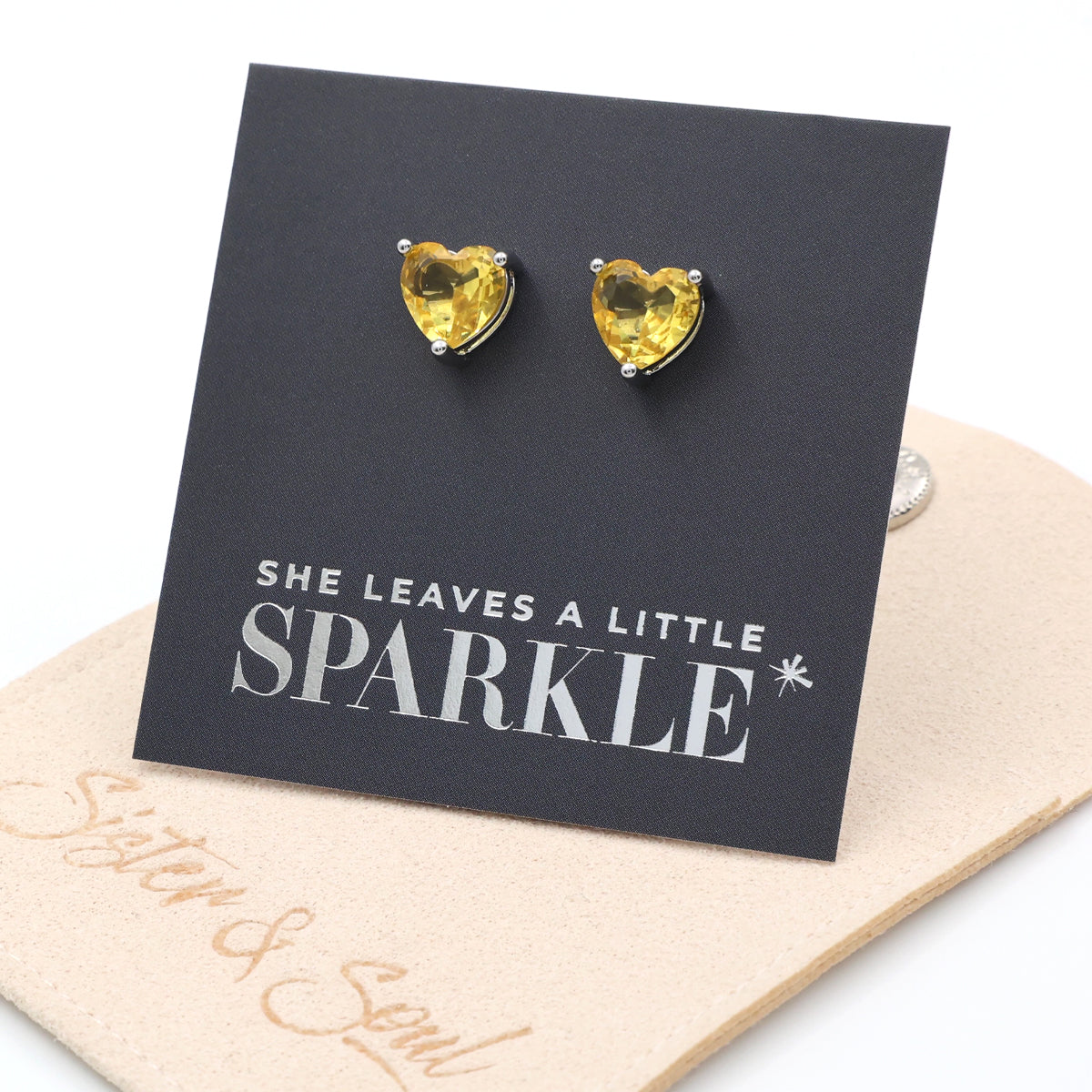 Heart-shaped yellow gemstone earrings on a card with 'She leaves a little sparkle' text.