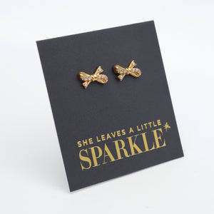 Gold bow shaped studs with clear gemstone features, on a card that says she leaves a little sparkle on a white background. 