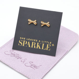 Gold bow shaped studs with clear gemstone features, on a card that says she leaves a little sparkle, and a purple gift pouch on a white background. 