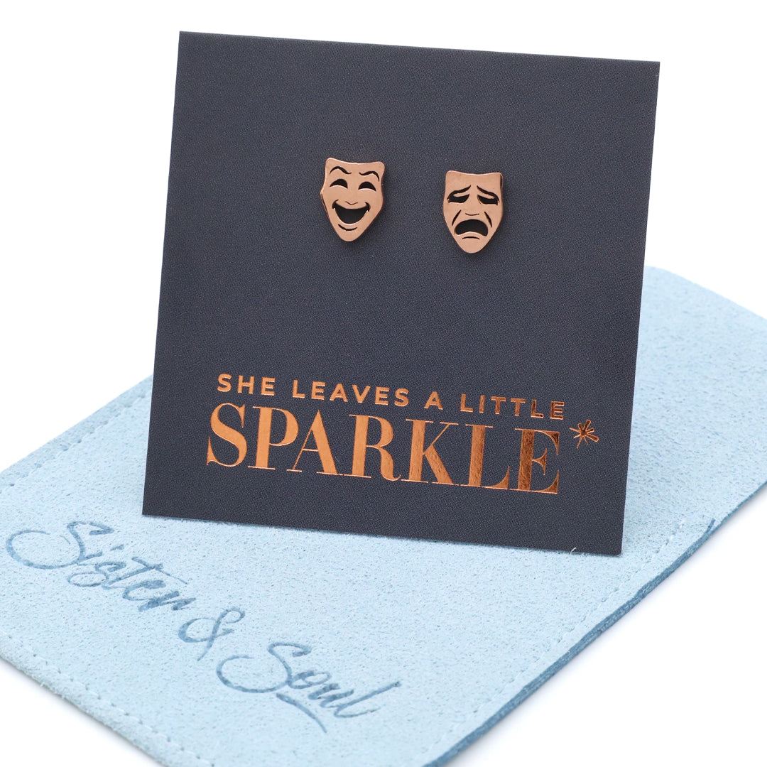 Rose gold theater mask earrings on a card with 'She leaves a little sparkle' text, displayed on a light blue fabric background.