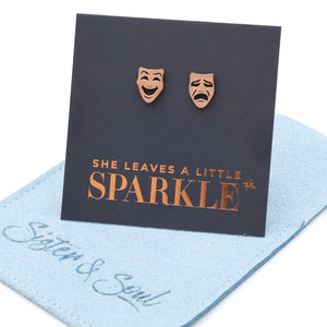 Rose gold theater mask earrings on a card with 'She leaves a little sparkle' text, displayed on a light blue fabric background.