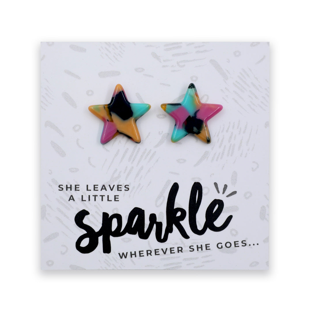 Two star-shaped stud earrings with a colorful marbled pattern in pink, blue, yellow, and black, displayed on a white printed card with the text 'SHE LEAVES A LITTLE sparkle WHEREVER SHE GOES.