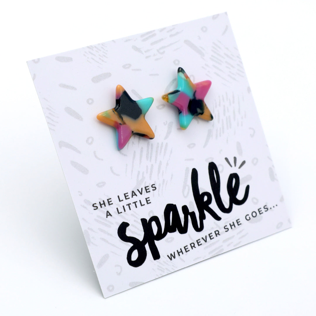 Two star-shaped stud earrings with a multicolor marbled pattern of pink, teal, yellow, and black, displayed on a white printed card that reads 'SHE LEAVES A LITTLE sparkle WHEREVER SHE GOES.