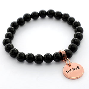 Black beaded bracelet with a rose gold 'BRAVE' charm on a white background