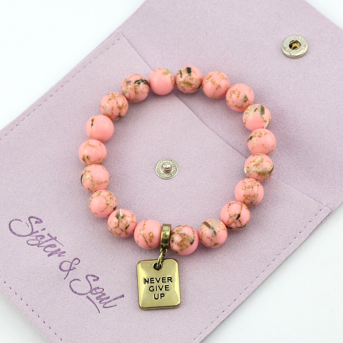 Pink beaded bracelet with 'Never Give Up' charm on a mauve pouch, featuring the brand 'Sister & Soul'.
