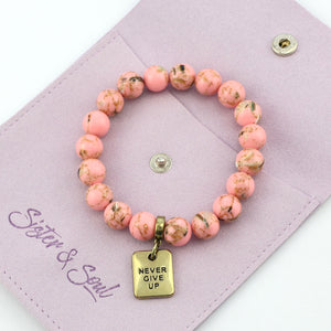 Pink beaded bracelet with 'Never Give Up' charm on a mauve pouch, featuring the brand 'Sister & Soul'.