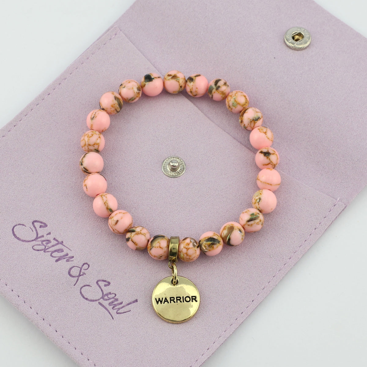 Pink beaded bracelet with a 'Warrior' charm on a mauve pouch with 'Sister & Soul' branding.