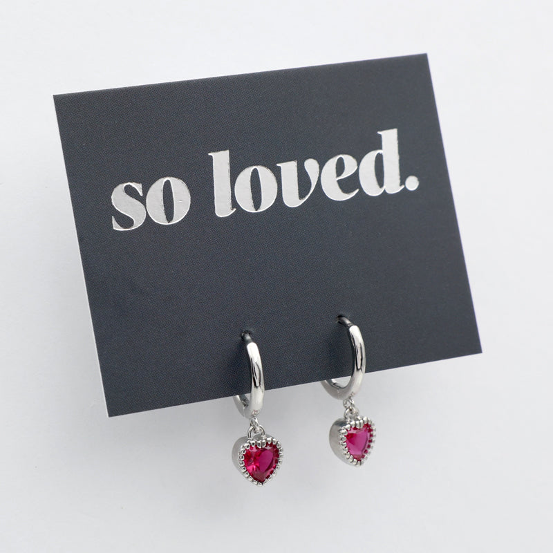 Pink heart shaped huggies dangle earrings presented on a grey card with silver 'so loved' writing.