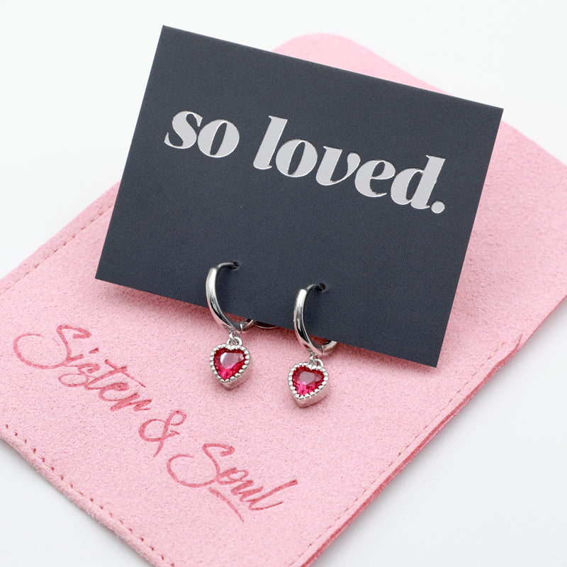 Pink heart shaped huggies dangle earrings presented on a grey card with silver 'so loved' writing sitting on a pink 'Sister & Soul' pouch.